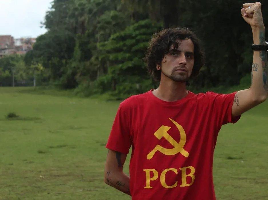 Giovani Damico, candidato ao governo da Bahia pelo PCB. Ele é branco, tem cabelo cahceado e escuro e levanta o braço com punho cerrado. Atrás, uma floresta - Metrópoles