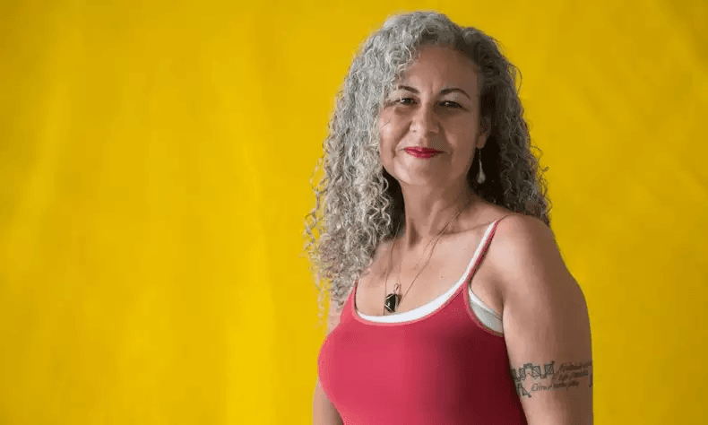 Lorene Figueiredo, candidata ao governo de Minas Gerais pelo Psol. Ela é parda, tem cabelo longo, cacheado e grisalho e olha para a câmera, sorrindo, sob fundo amarelo - Metrópoles